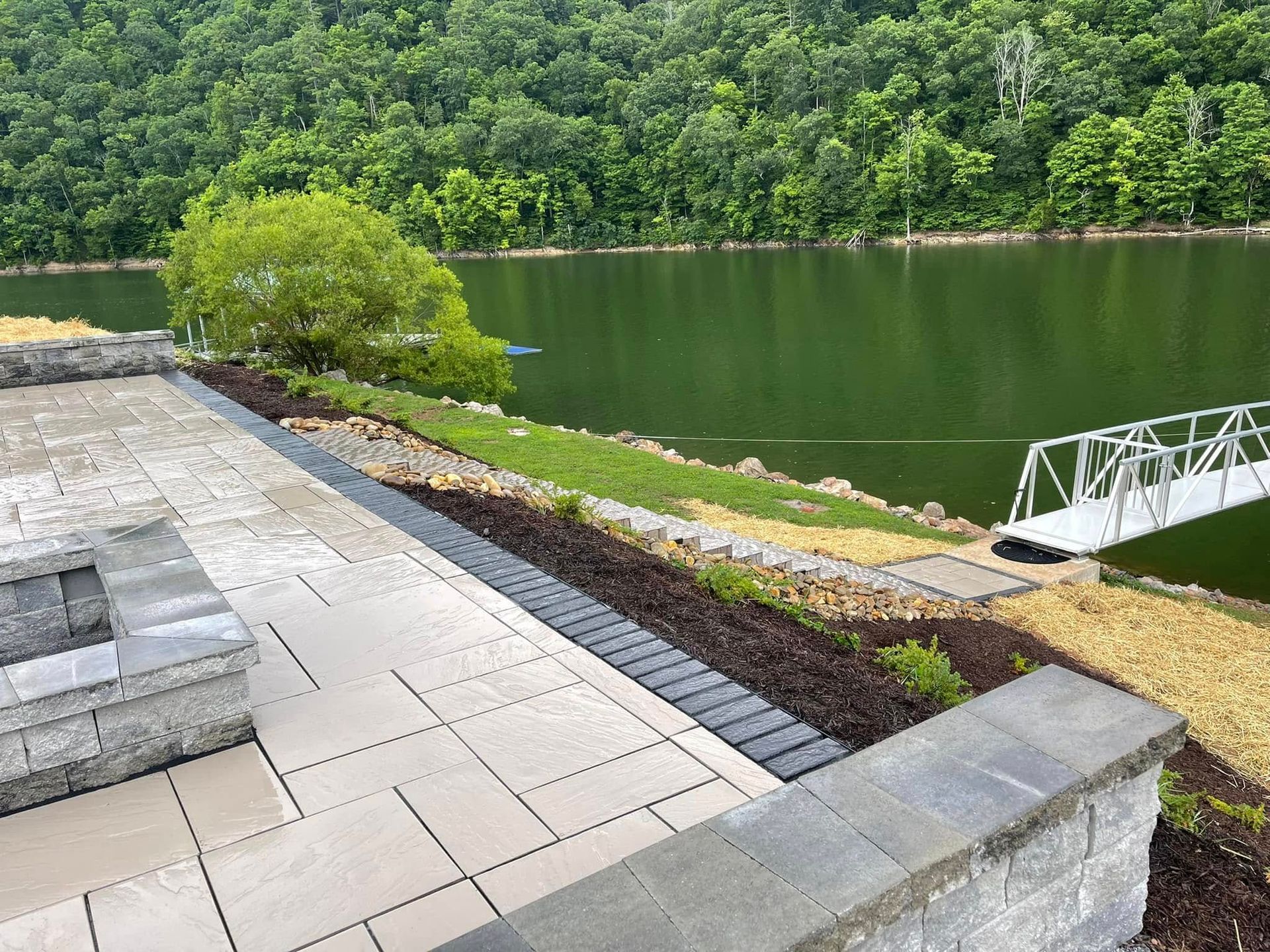 Paved patio beside a pond, with a raised square fire pit and surrounding garden beds