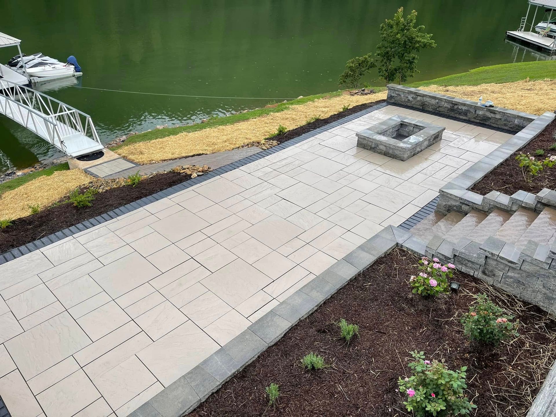 Paved patio beside a pond, with a raised fire pit and landscaped garden beds.