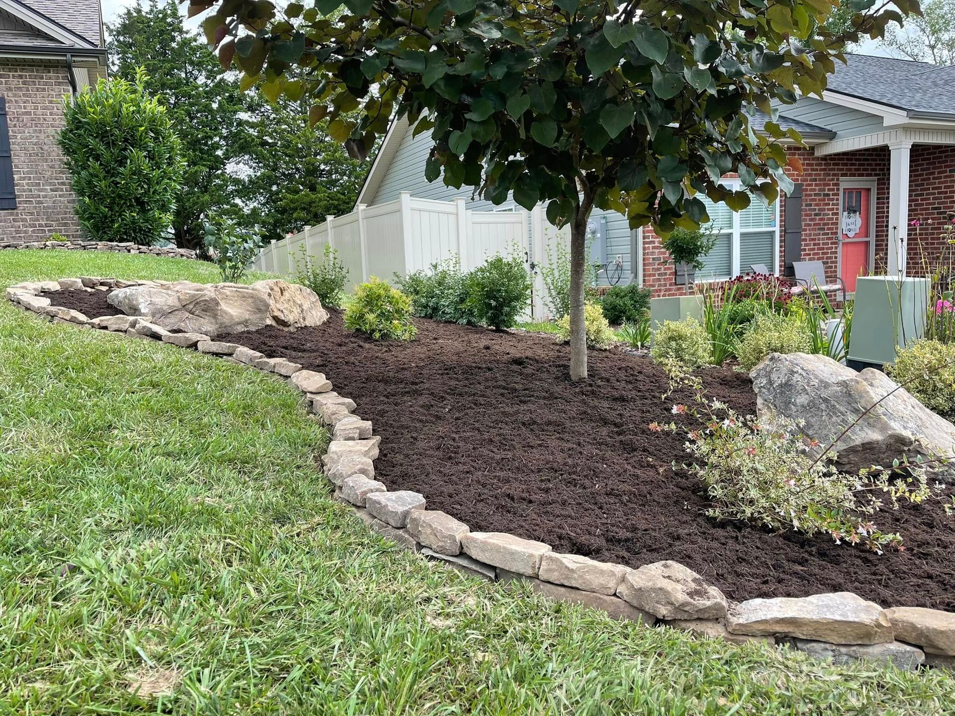 Curved mulched garden bed with stone edging and a tree beside a house front yard.