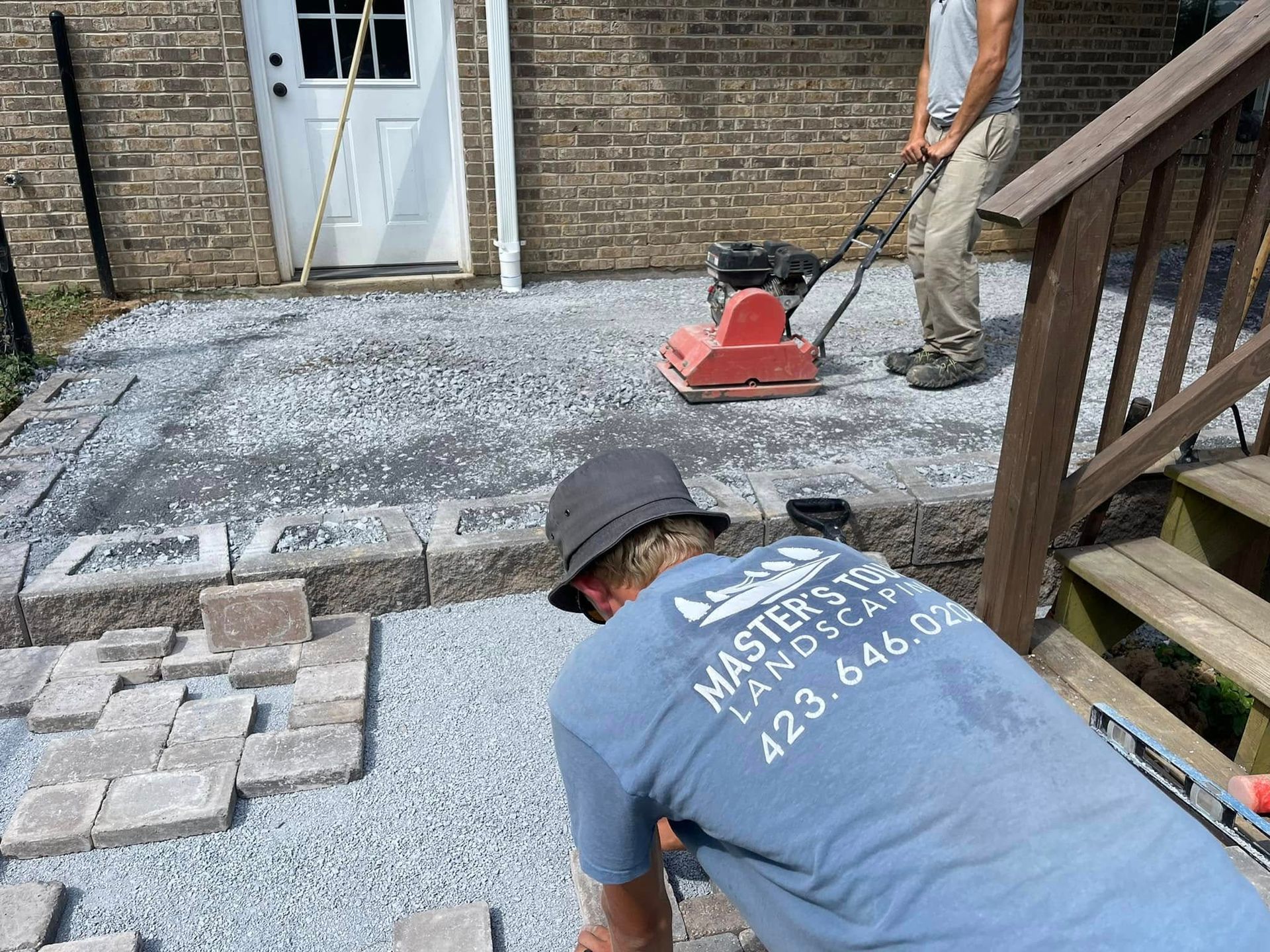 Workers installing a brick patio beside a house, with pavers, a compactor, and stairs visible.