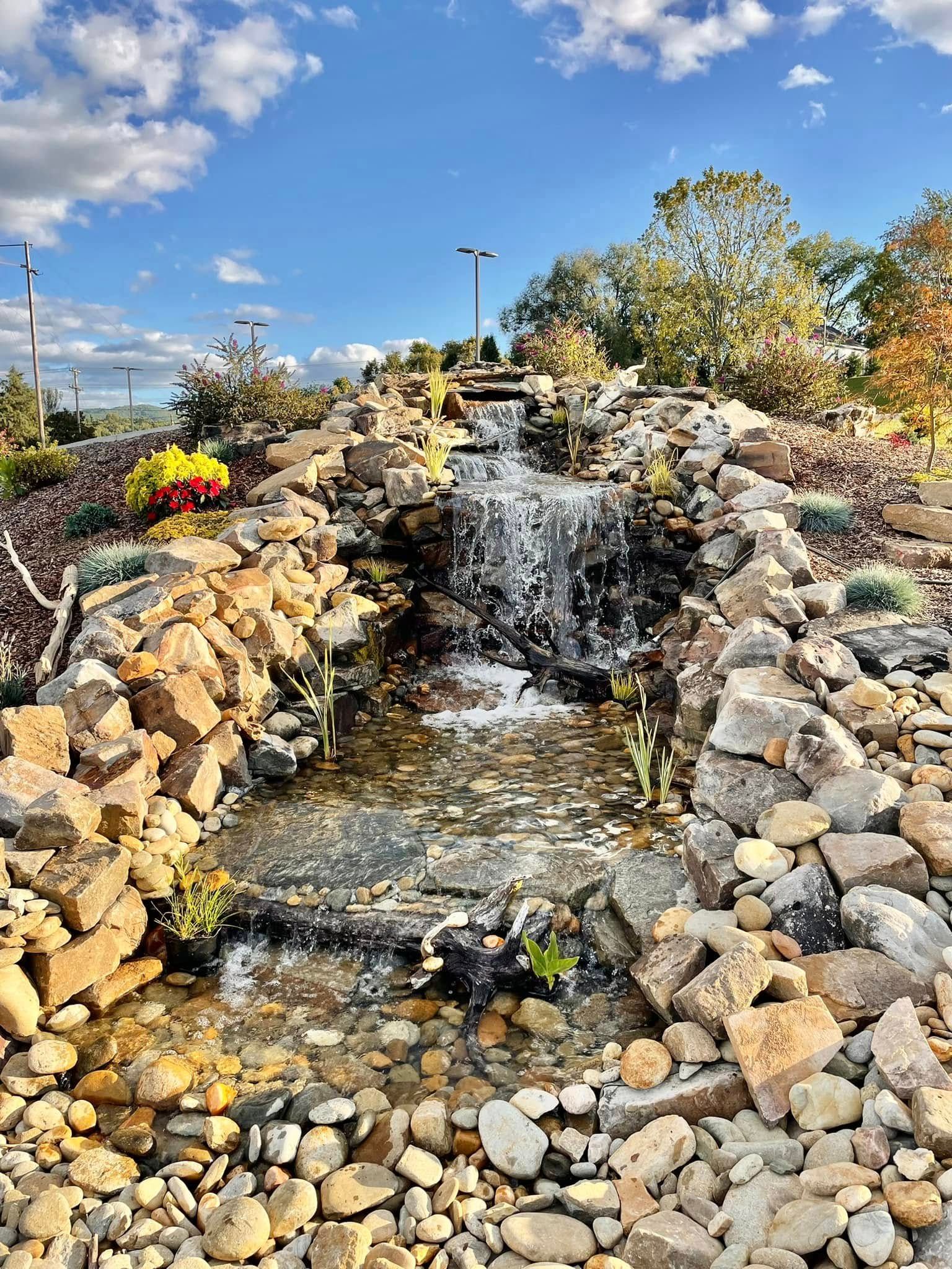 Rock waterfall cascading into a stone-lined pond with colorful flowers and trees in the background