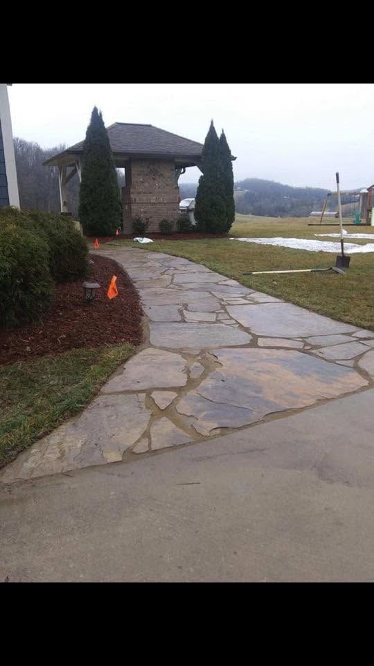 Stone walkway curving through a landscaped yard beside a house with tall evergreen trees and mountains in the distance