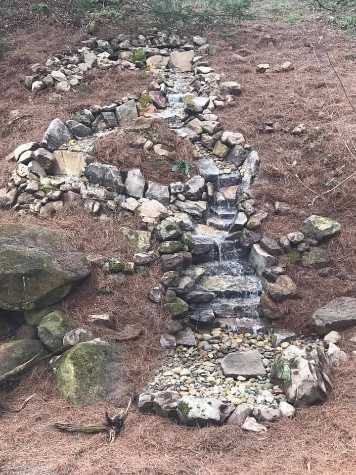 Stone-lined garden waterfall trickling down a bare, brown hillside.