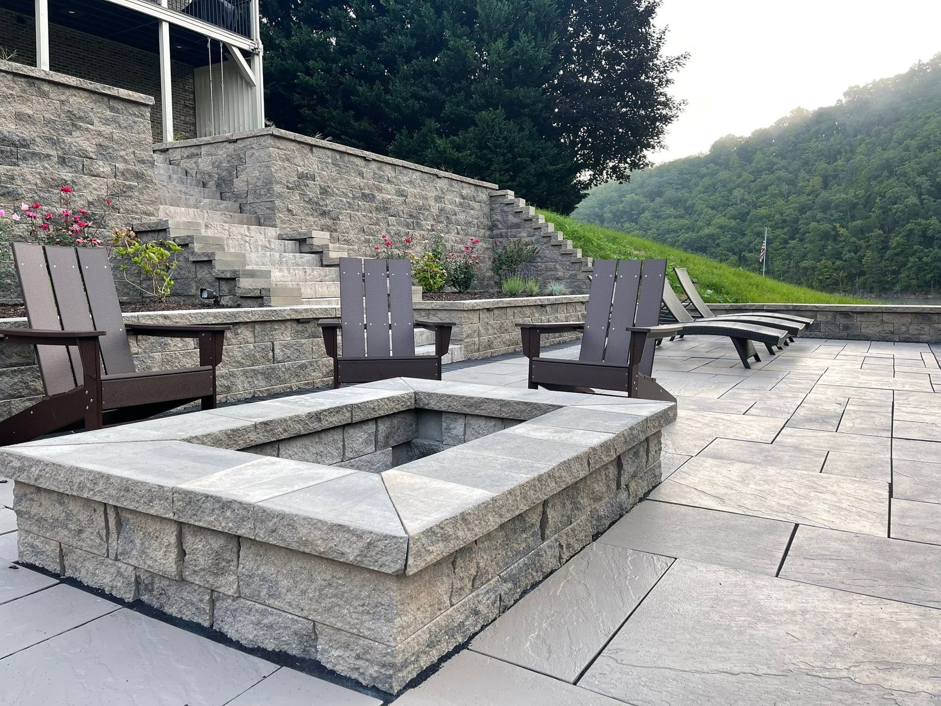 Stone fire pit with Adirondack chairs on a paved patio beside steps and a grassy hillside
