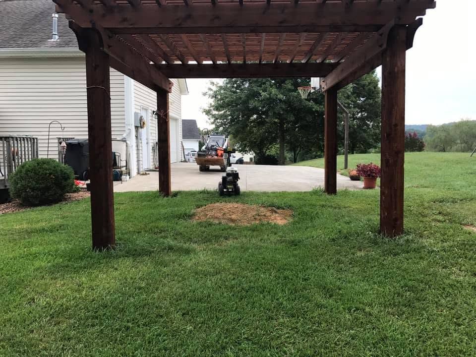 Wooden pergola over a grassy yard beside a house, with a small cart centered underneath.
