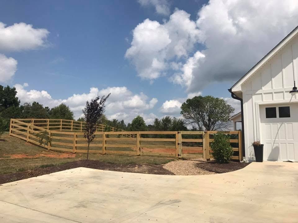 White barn beside a paved drive with wooden fence, small trees, and open fields under a cloudy blue sky