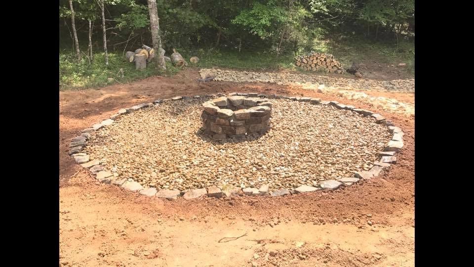 Circular stone fire pit in a gravel ring, surrounded by red dirt and forest greenery.