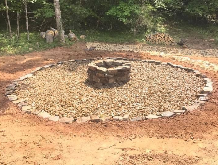 Stone fire pit in a circular gravel area outdoors, surrounded by dirt and trees.
