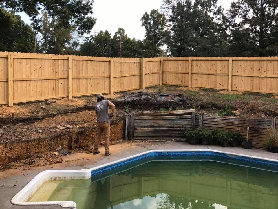 Person standing by an empty backyard pool beside a new wooden fence and dirt-filled landscaping.