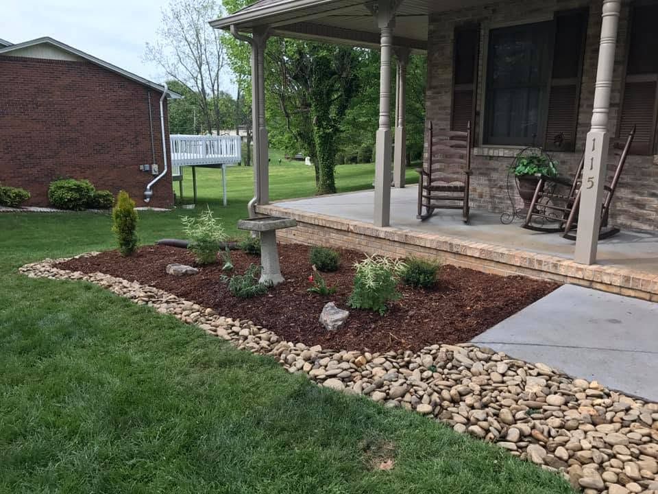 Front porch with wooden rocking chairs, stone border, mulch bed, and a small birdbath beside a lawn