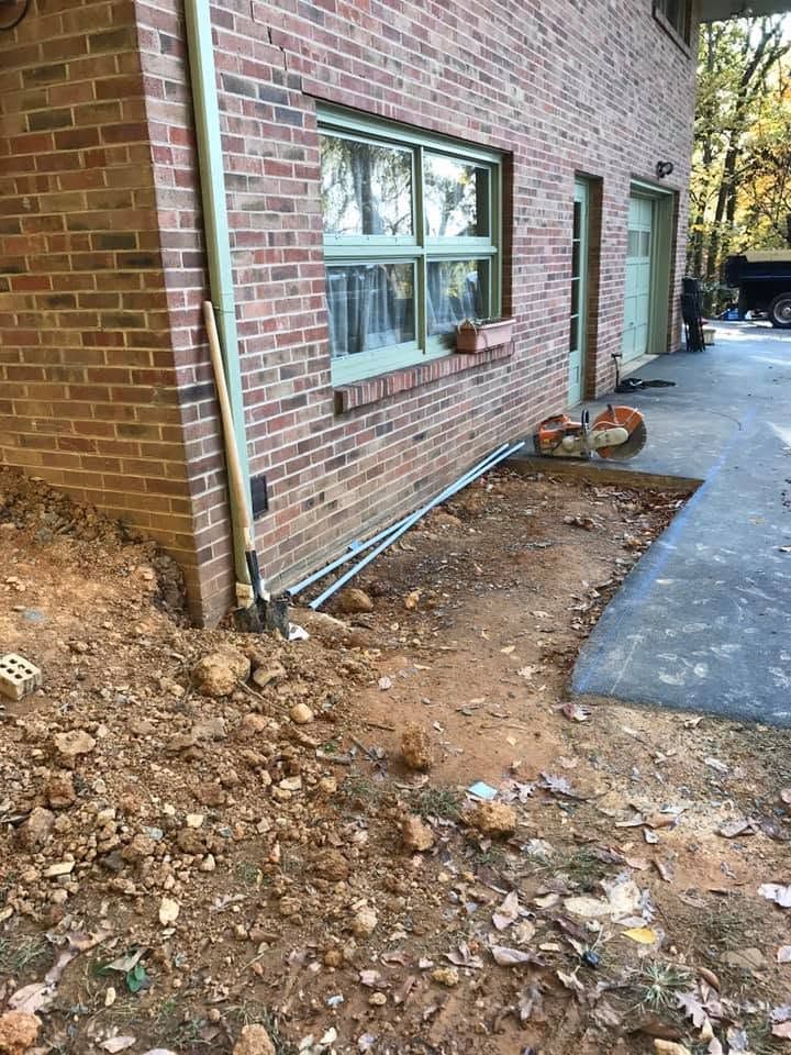 Brick house side with windows, exposed dirt trench, and a paved walkway along the wall.