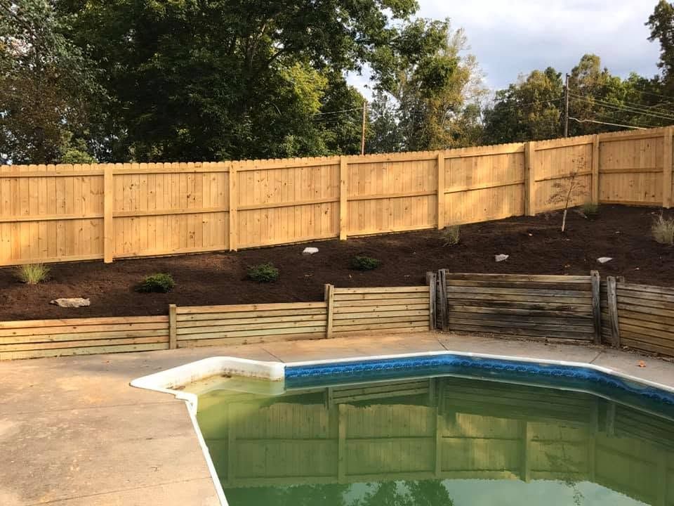Backyard pool with green water, wooden fence, mulch hillside, and trees in the background