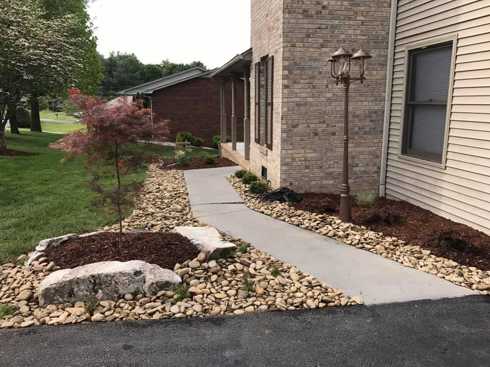 Concrete walkway beside a brick house with rock landscaping and shrubs in a suburban yard