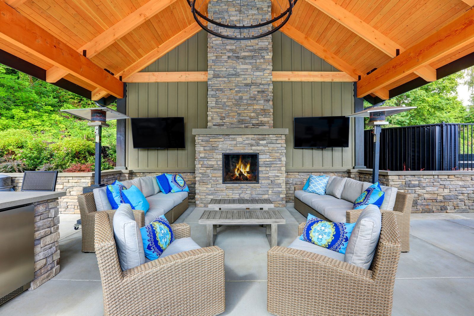 Covered outdoor patio with wicker seating, blue cushions, and a stone fireplace under a wooden pavilion.