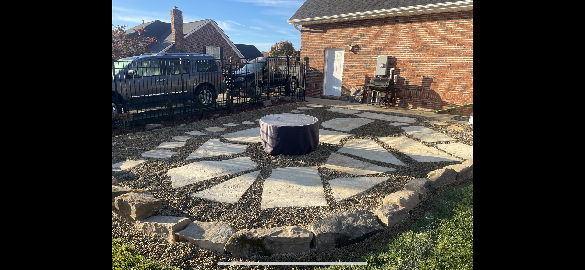 Backyard stone fire pit with a central black metal stump, beside a brick wall and parked van