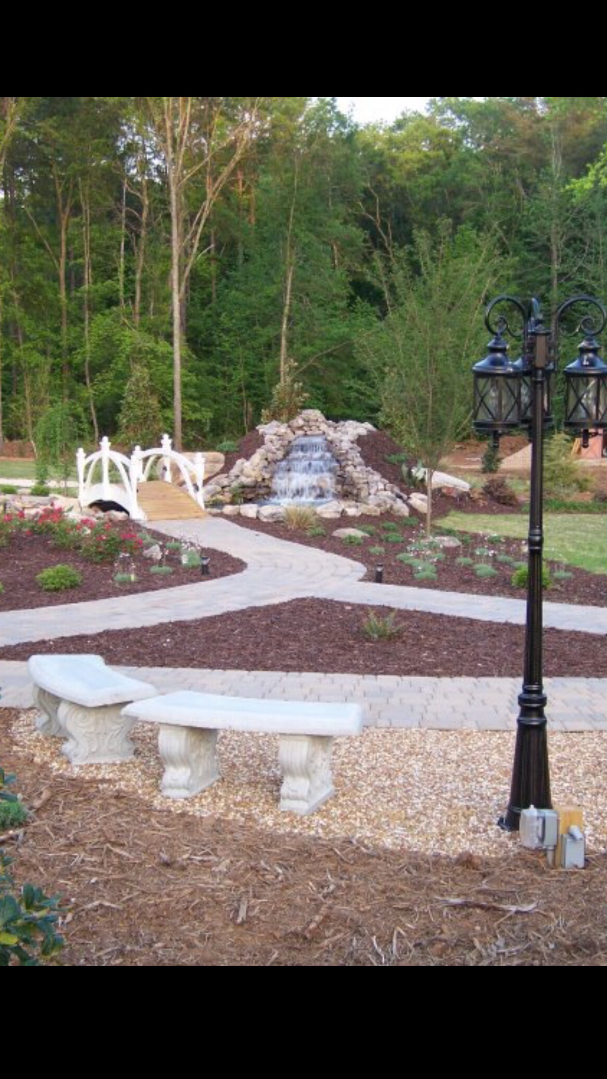 Garden path winding past white benches, a small waterfall, and a black lamp post among trees.
