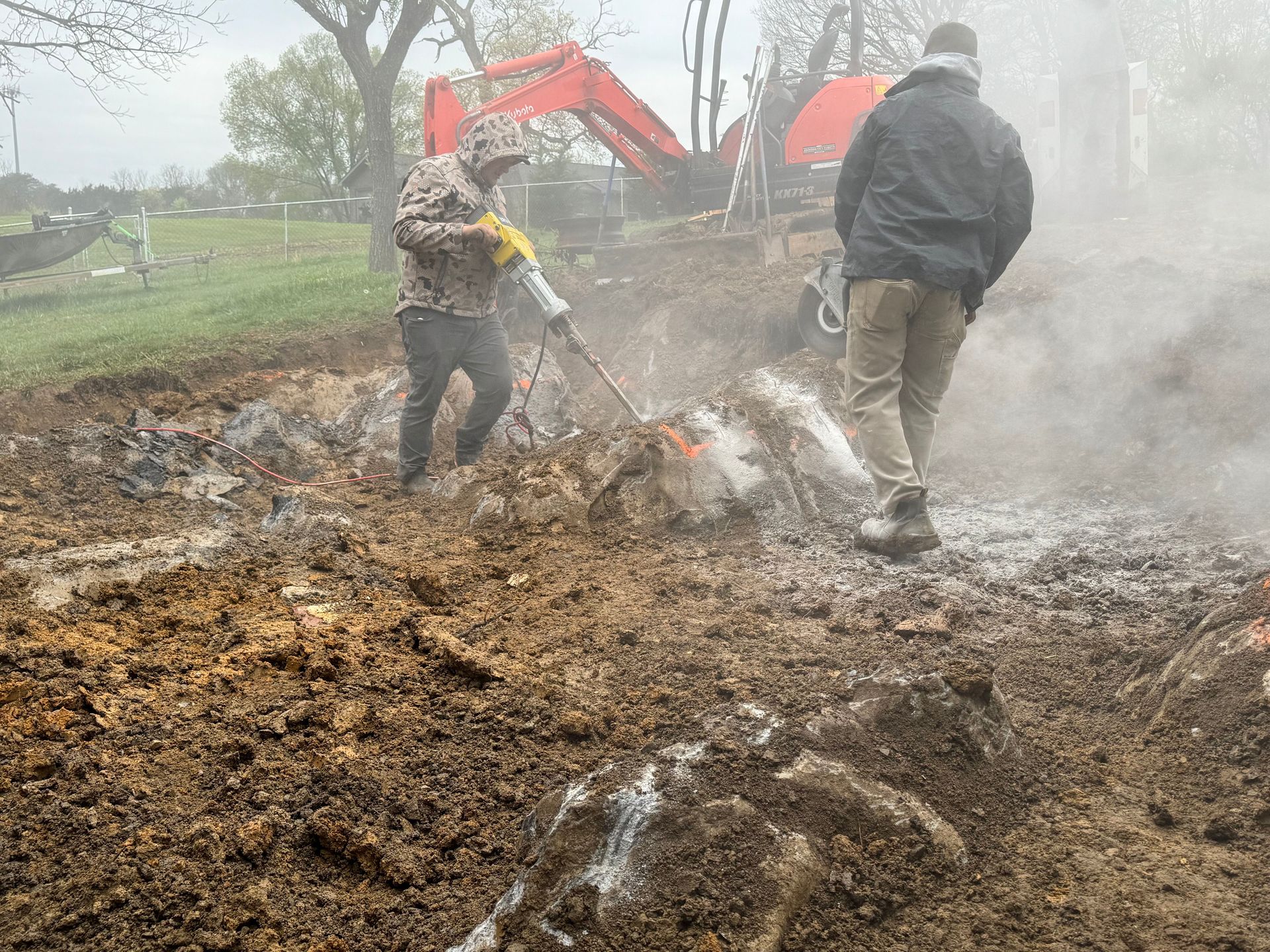 Two workers spraying water on a smoky muddy excavation site with an excavator in the background
