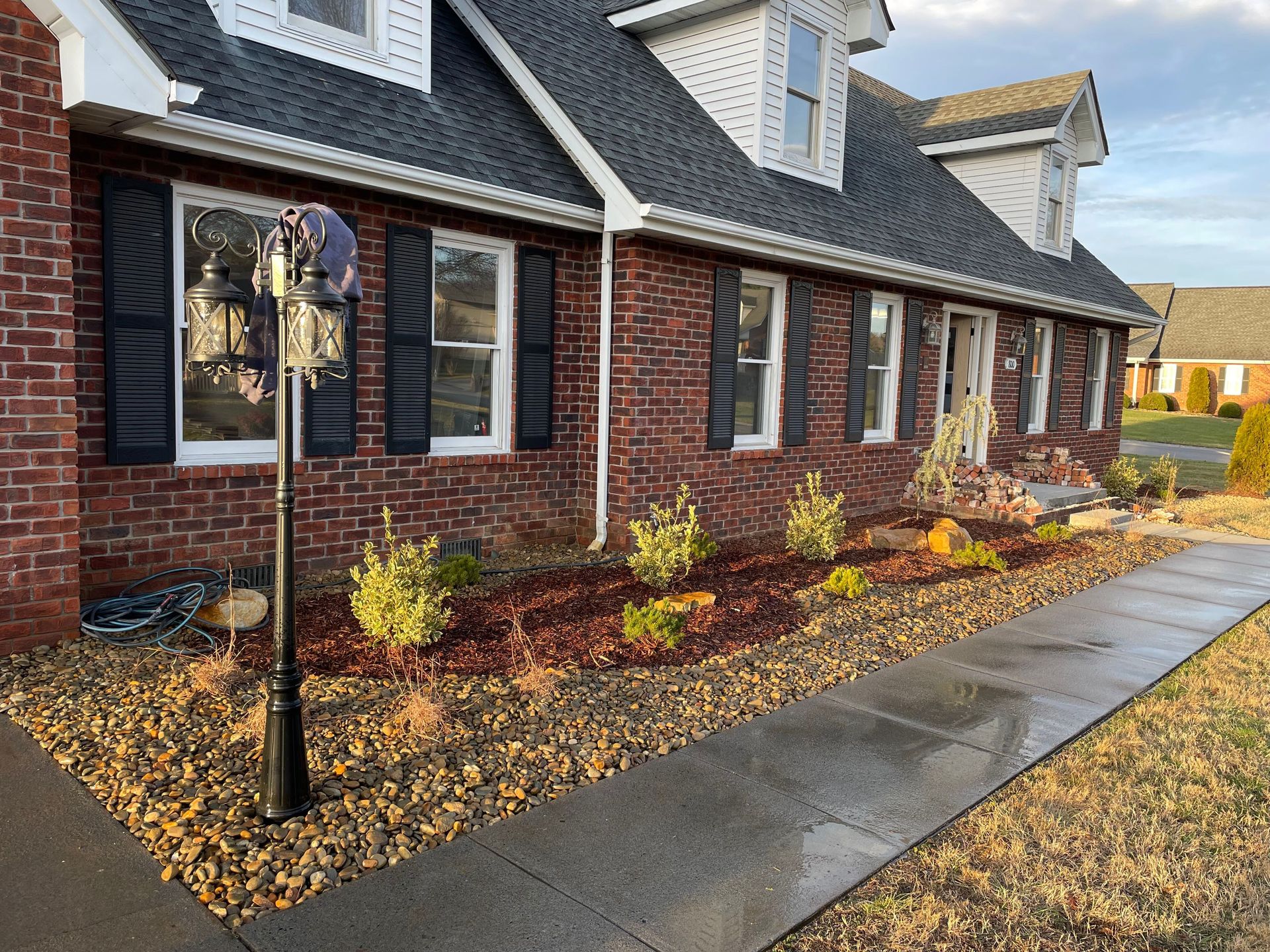 Brick house front with black shutters, landscaped mulch bed, and wet sidewalk along the facade