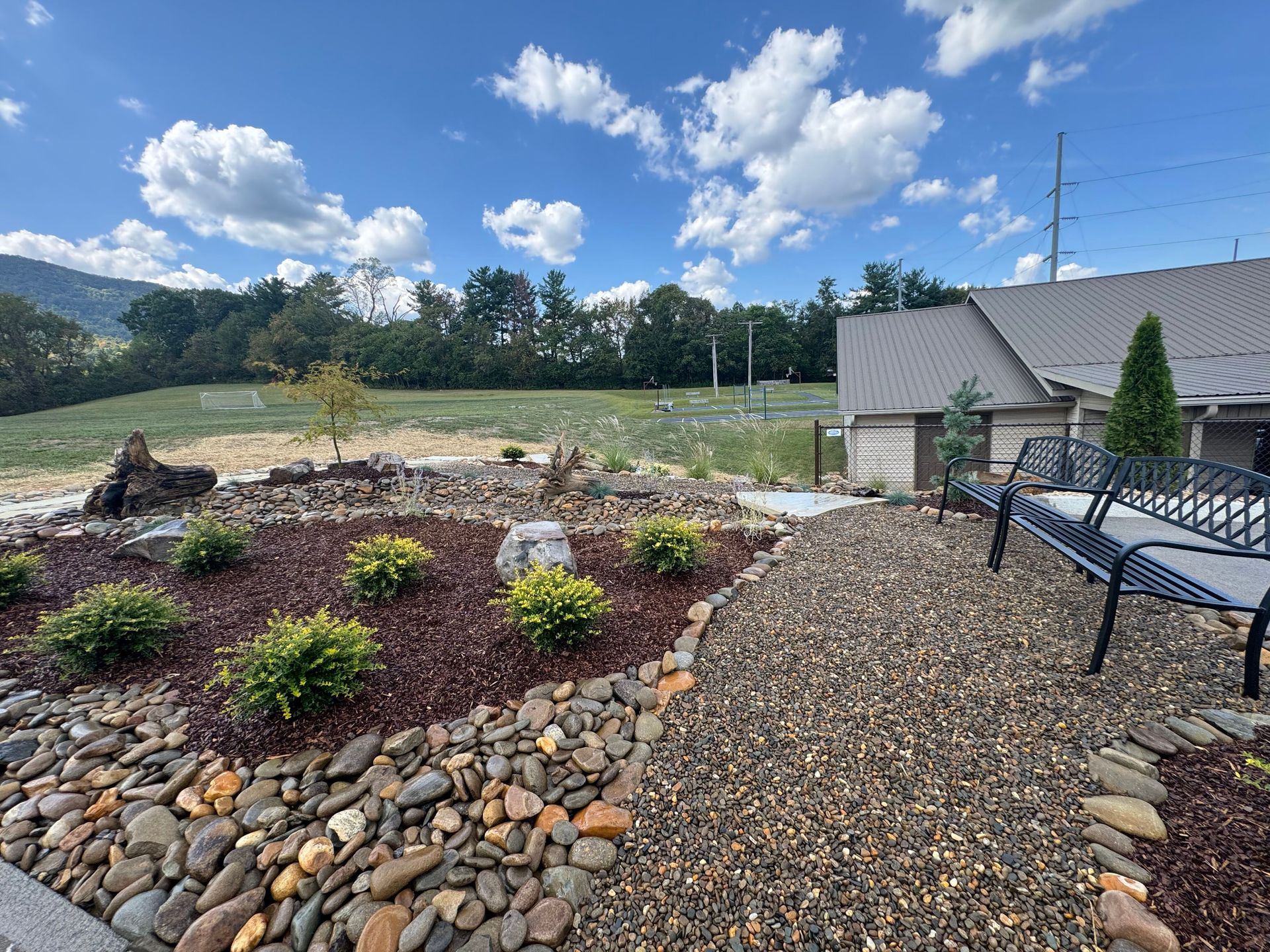 Landscaped backyard with rock beds, shrubs, a blue bench, and a small building under a bright cloudy sky