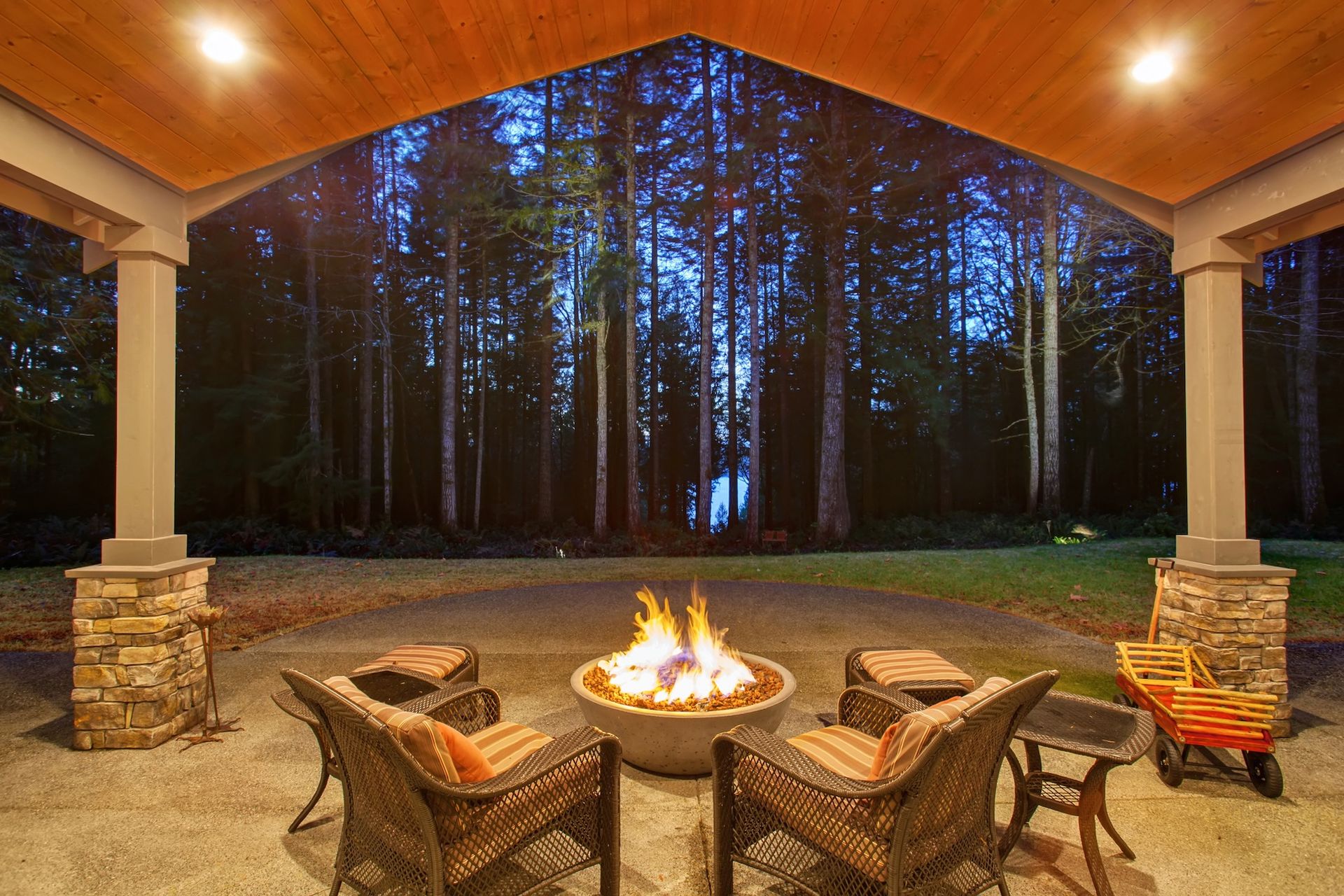 Covered patio with wicker chairs around a fire pit, overlooking a dark wooded forest at dusk.