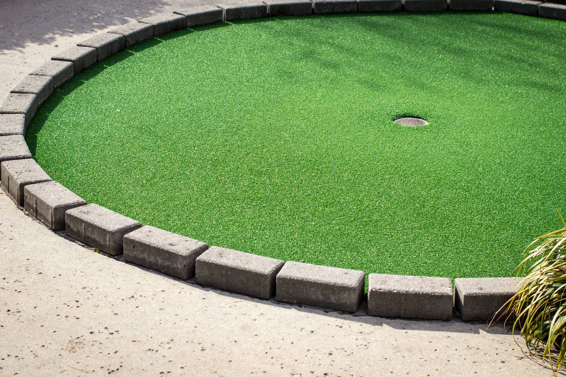 Circular green turf area bordered by light stone blocks beside a pale walkway