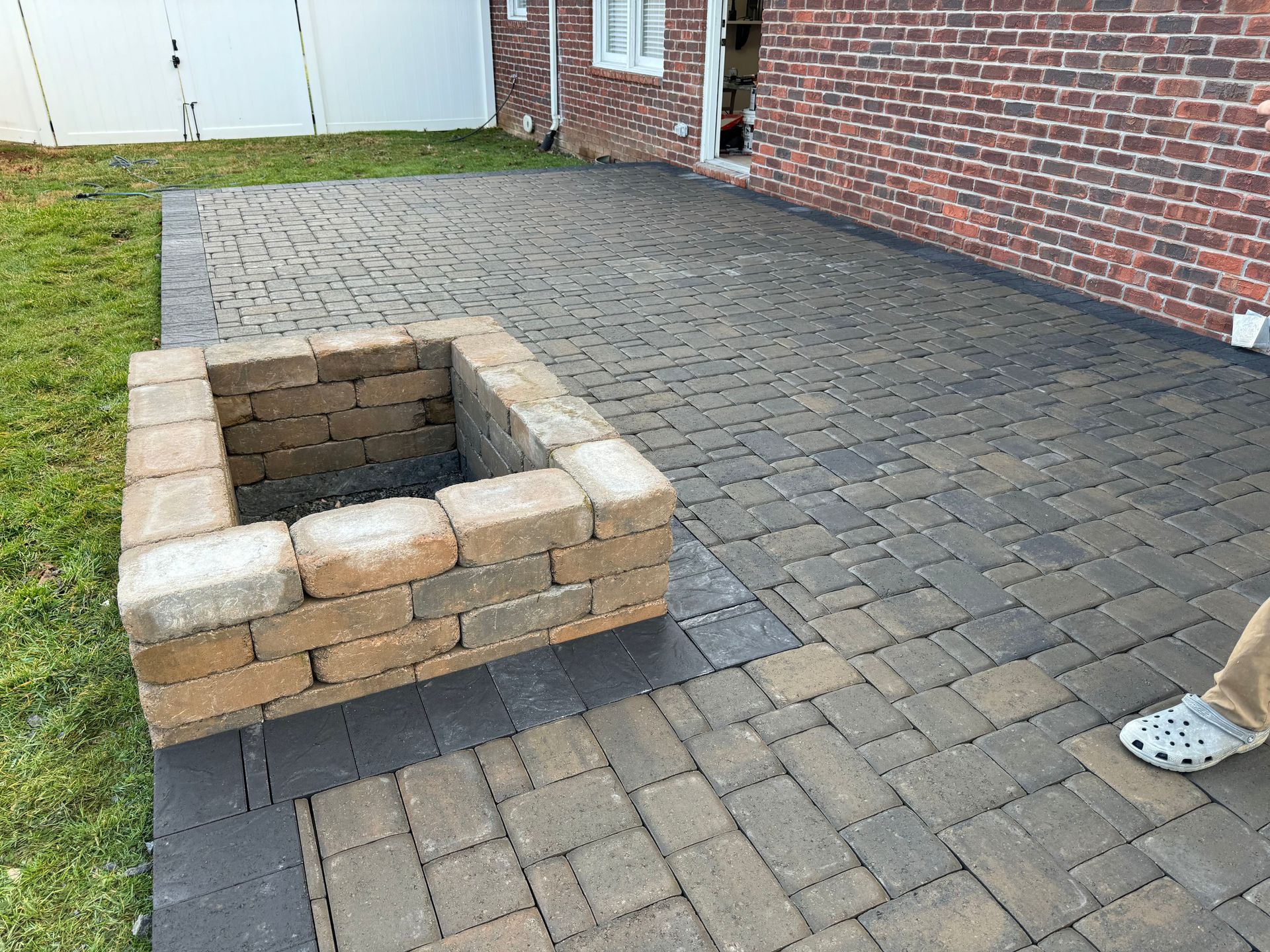 Brick fire pit on a paved patio beside a house, with part of a person’s shoe visible at right