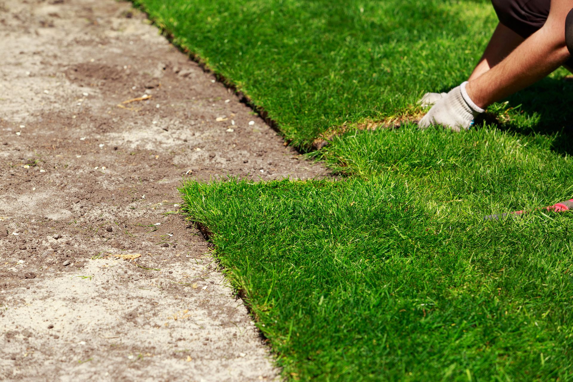 Person’s legs edging a green lawn with a garden tool beside a dirt path