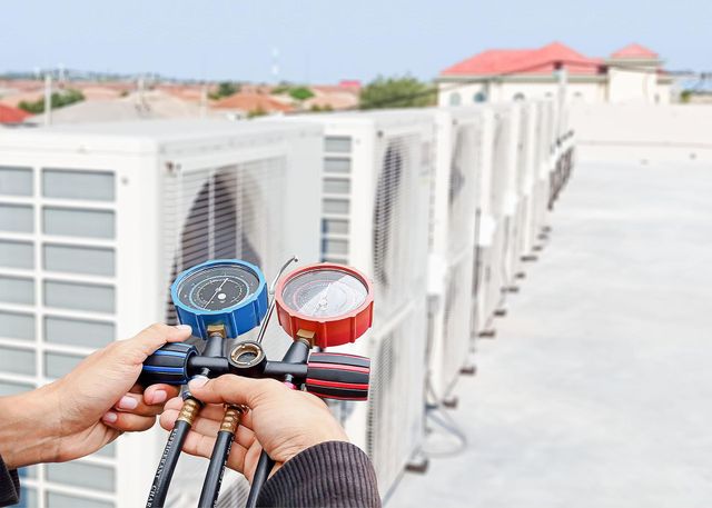 Hands holding blue and red manifold gauges connected to HVAC equipment on a rooftop.