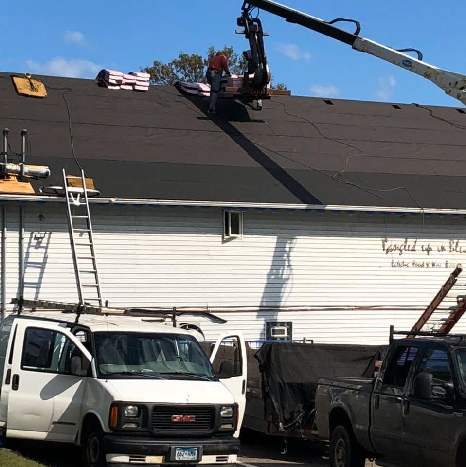 Roofers working on a dark roof with a crane, ladders, and work trucks below.