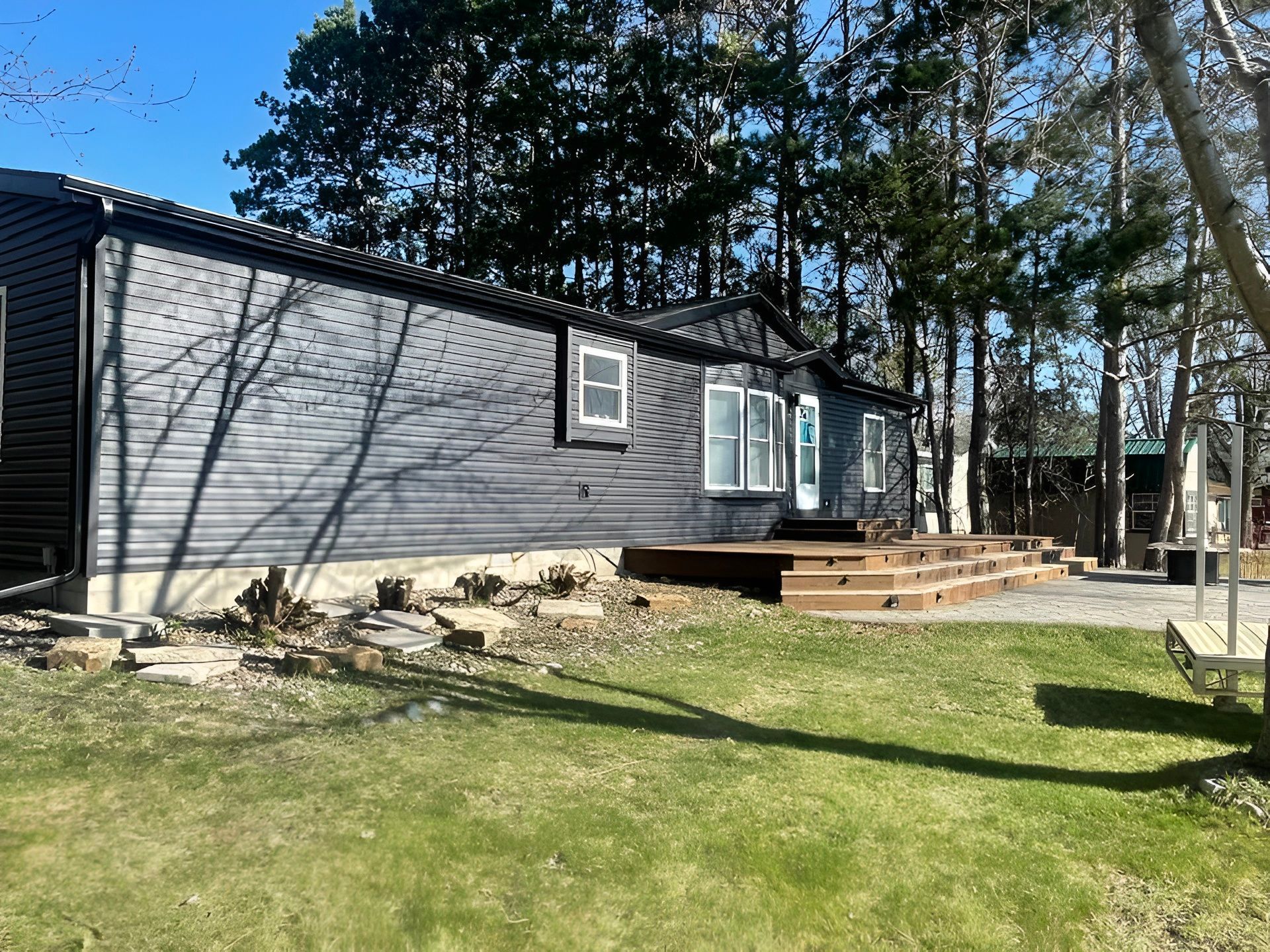 Gray house with a wooden deck and steps in a sunny yard, surrounded by trees