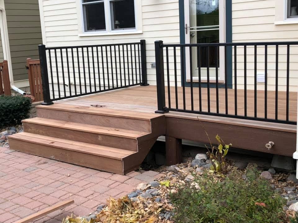 Wooden front steps with black railing leading to a house entry over a brick walkway.