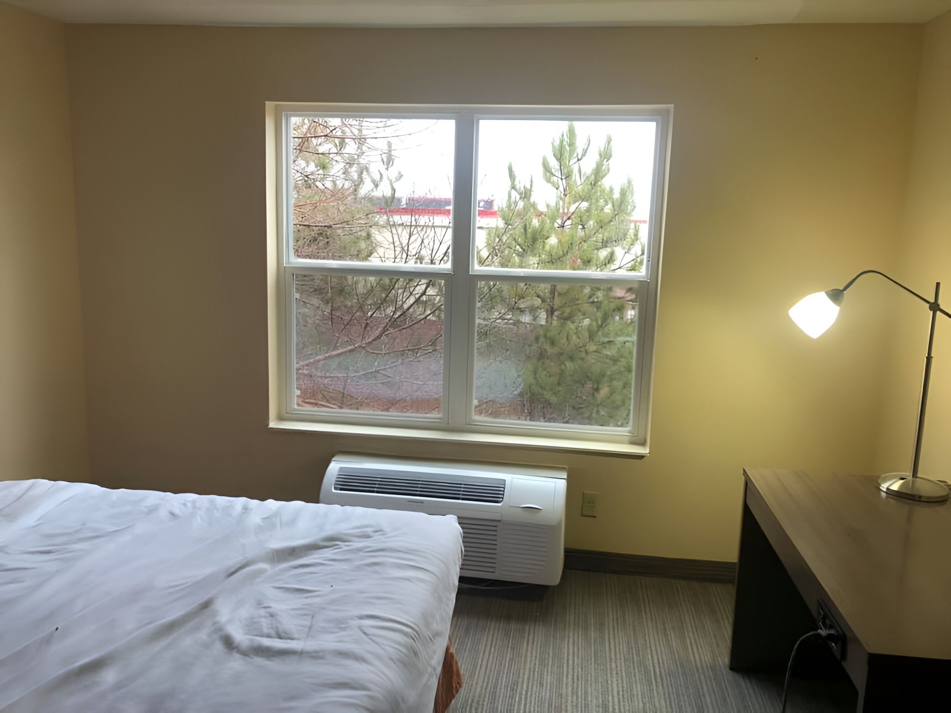 Bedroom with unmade bed, window, air conditioner, and lit desk lamp on a dark dresser.