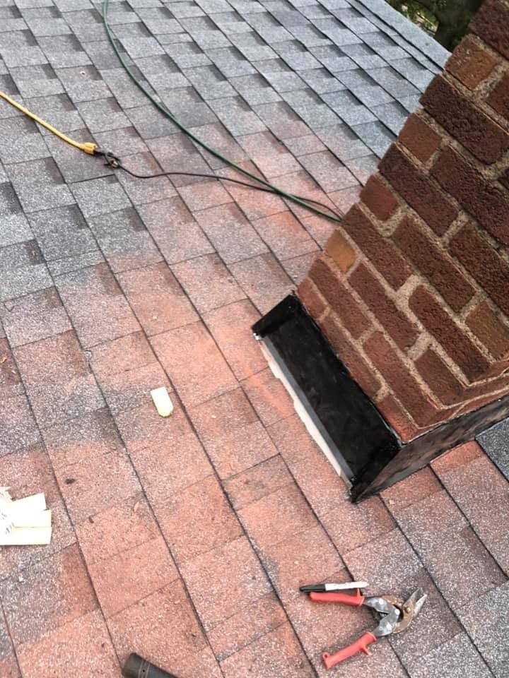 Roof shingles around a brick chimney with tools and a cord visible on the rooftop