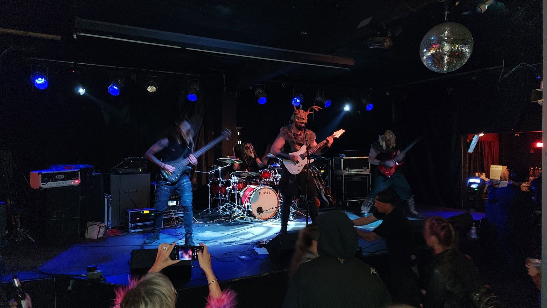 Live rock band performing on a dimly lit stage with blue lights and a disco ball, audience watching in front