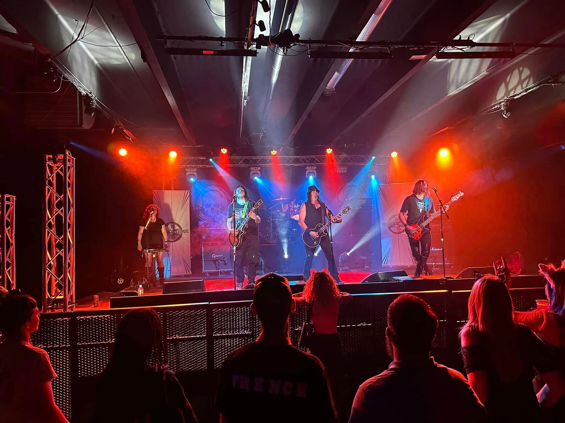 Live rock band performing onstage under red and blue lights, with audience silhouettes in front