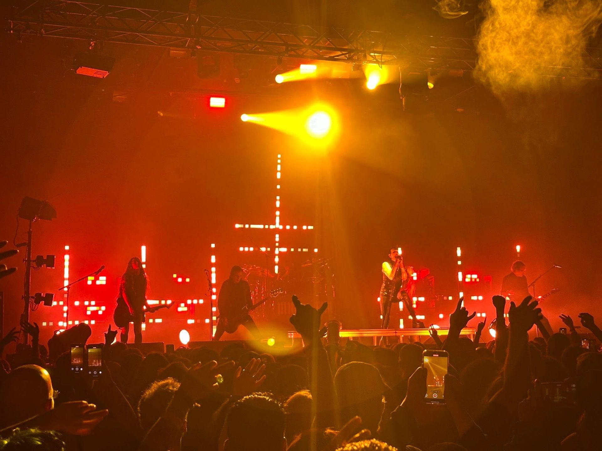 Concert stage lit with red and yellow lights, silhouetted performers, and a cheering crowd with raised hands