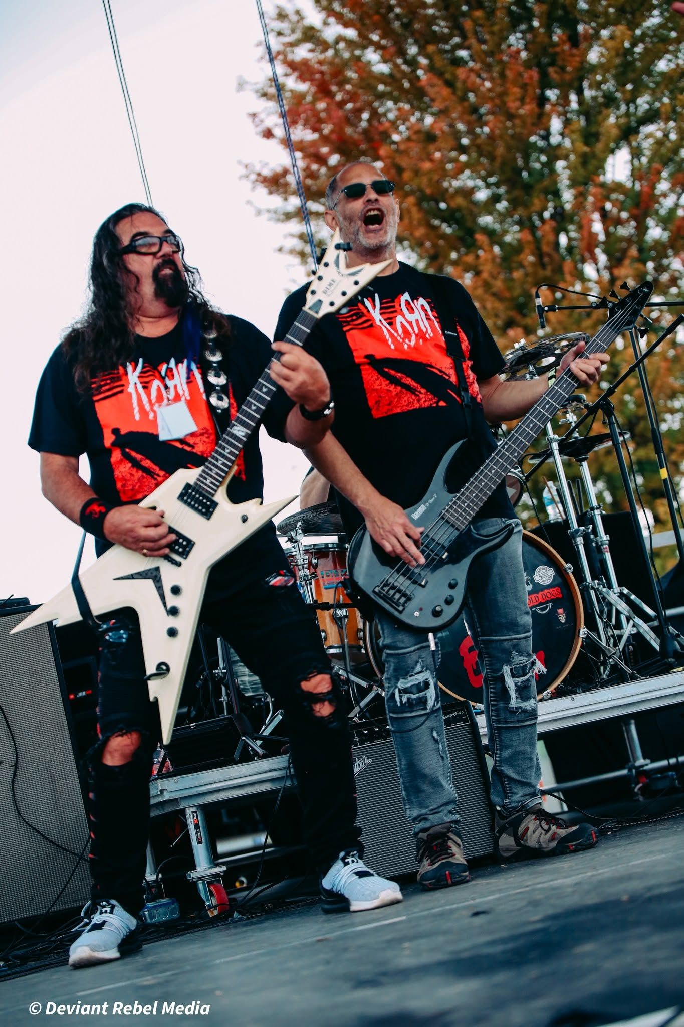 Two musicians playing electric guitars on an outdoor stage with autumn trees behind them
