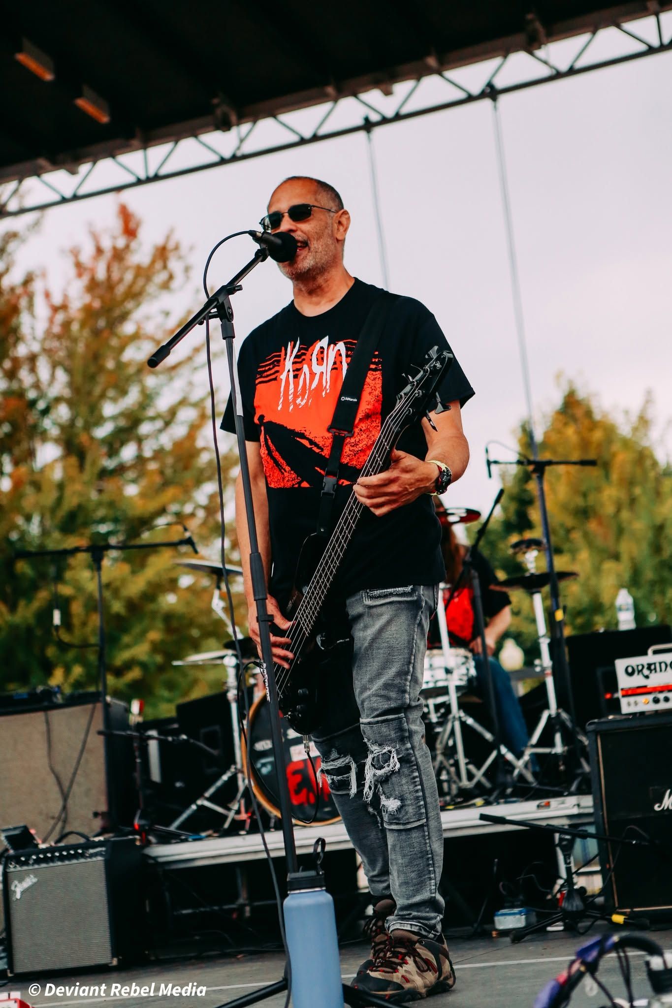 Singer with guitar on an outdoor stage, singing into a microphone in a black T-shirt and sunglasses