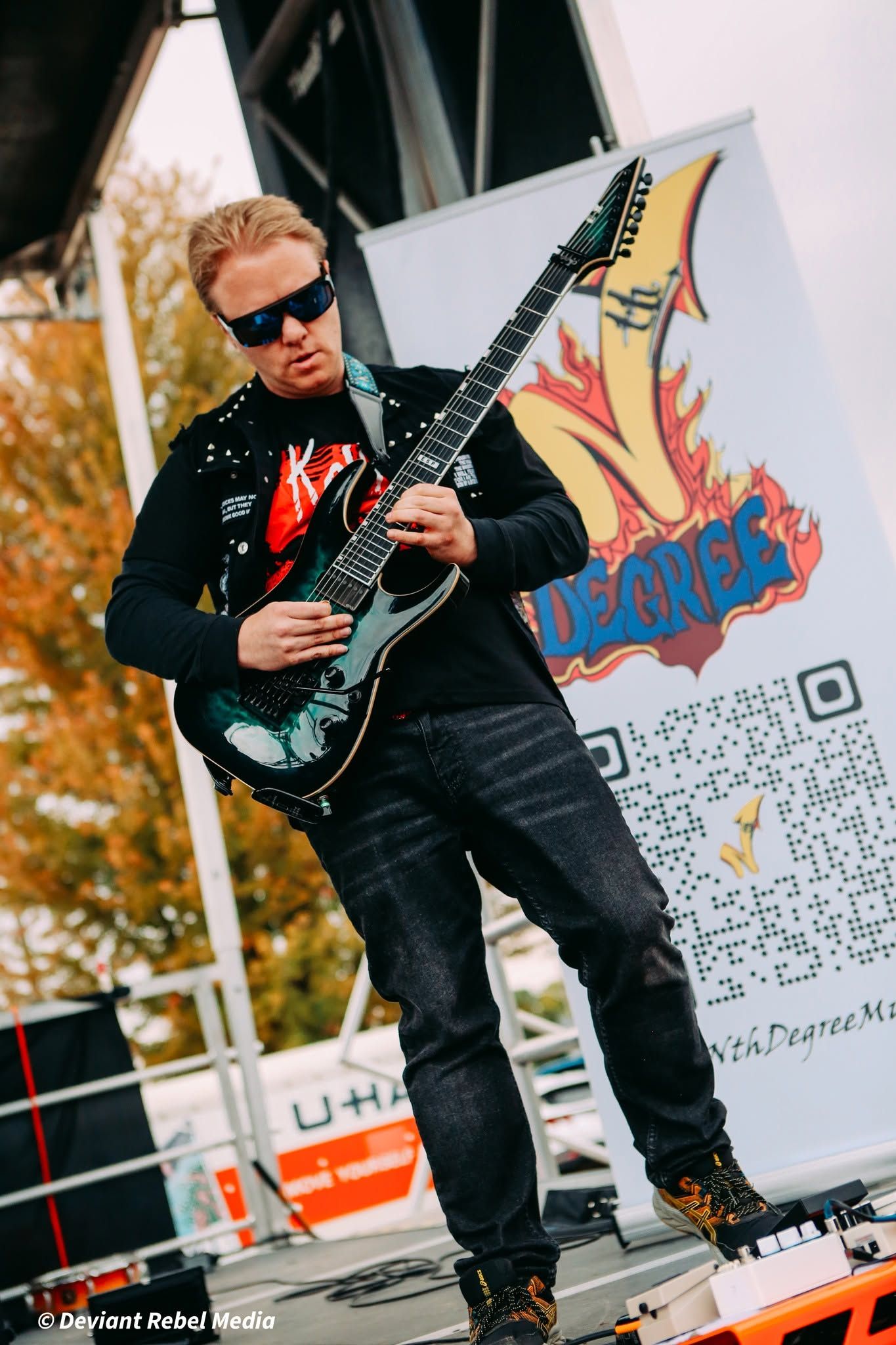 Musician playing electric guitar on an outdoor stage beside a banner and QR code