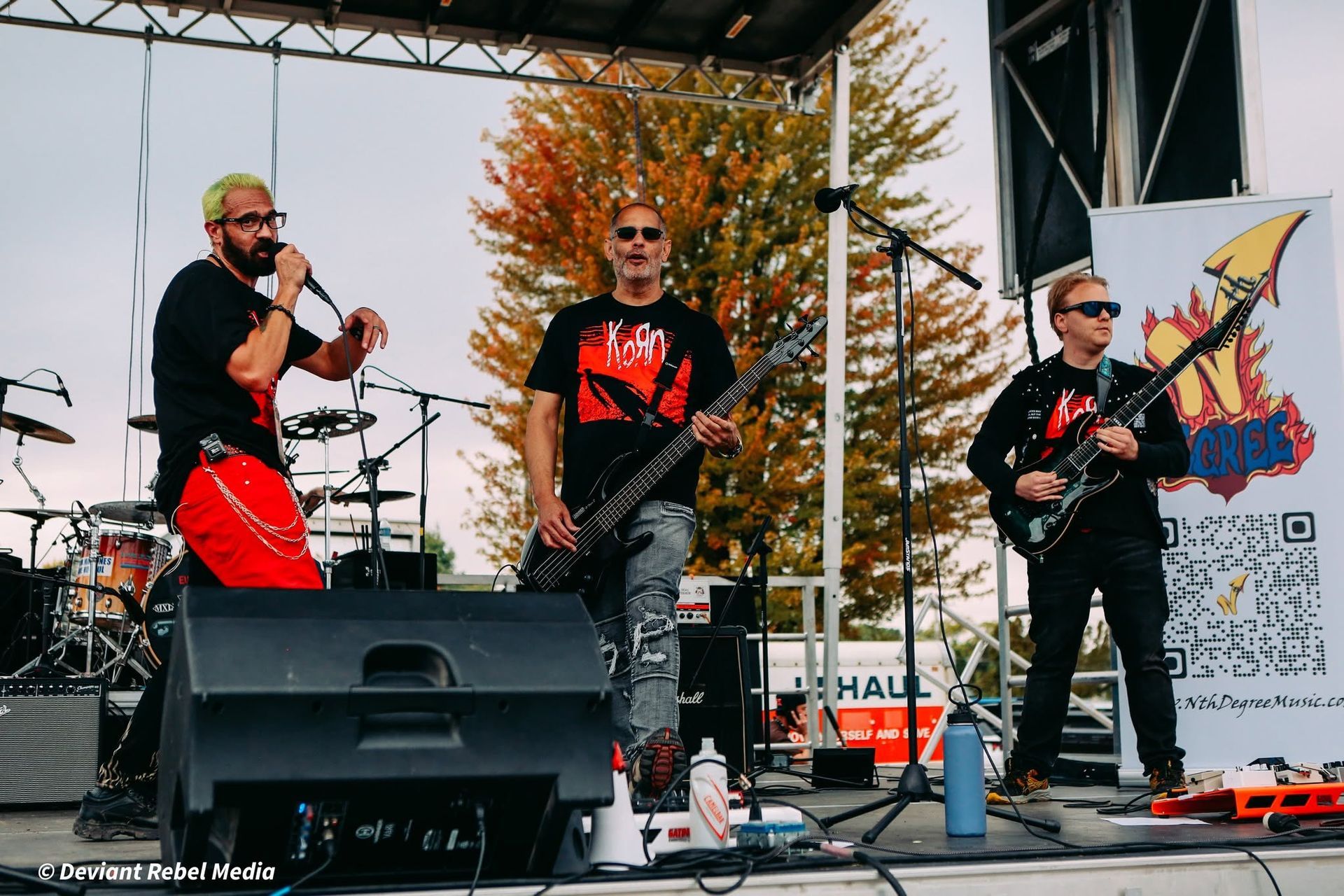 Three musicians performing on an outdoor stage, with drums, guitars, and a banner backdrop.