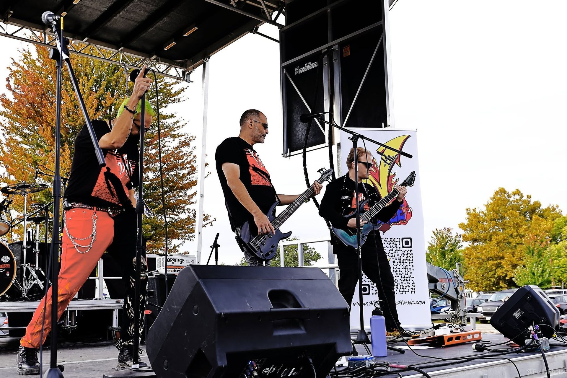 Band performing on an outdoor stage with guitars and microphones, autumn trees in the background