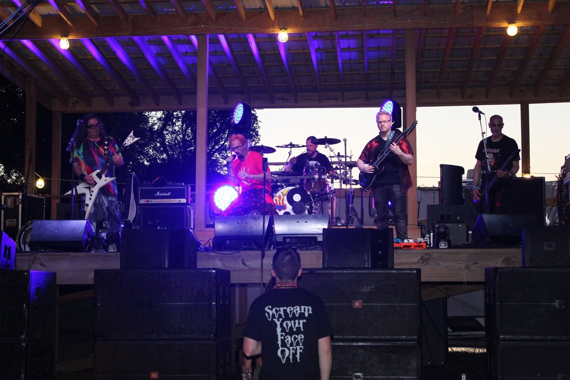 Live band performing on an outdoor stage under purple lights, with drums and guitars at dusk.
