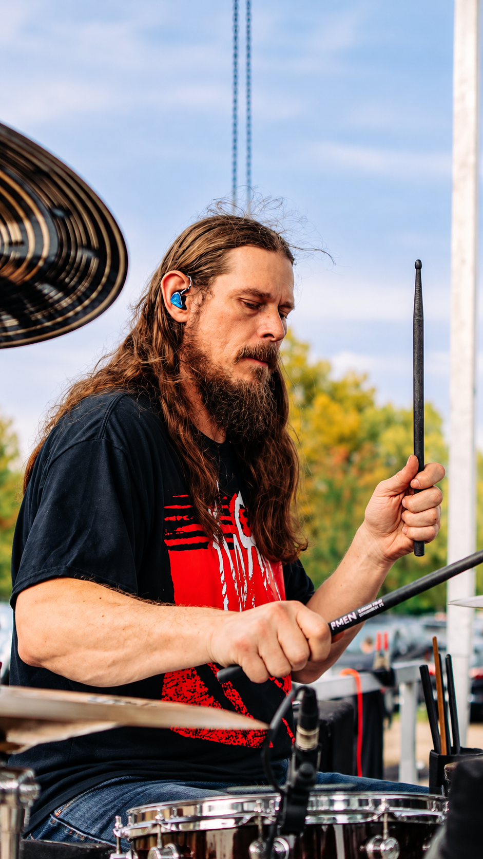 Long-haired drummer playing outdoors with a black shirt and red graphic tee, holding drumsticks.