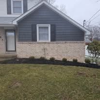Exterior of a house with dark blue siding above a brick base, black mulch and green lawn.