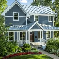 Blue house with white trim and metal roof, surrounded by green shrubs and flowers.