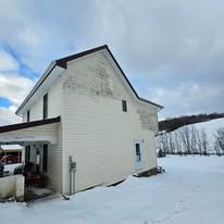 Beige two-story house with faded siding, snowy field, and overcast sky.
