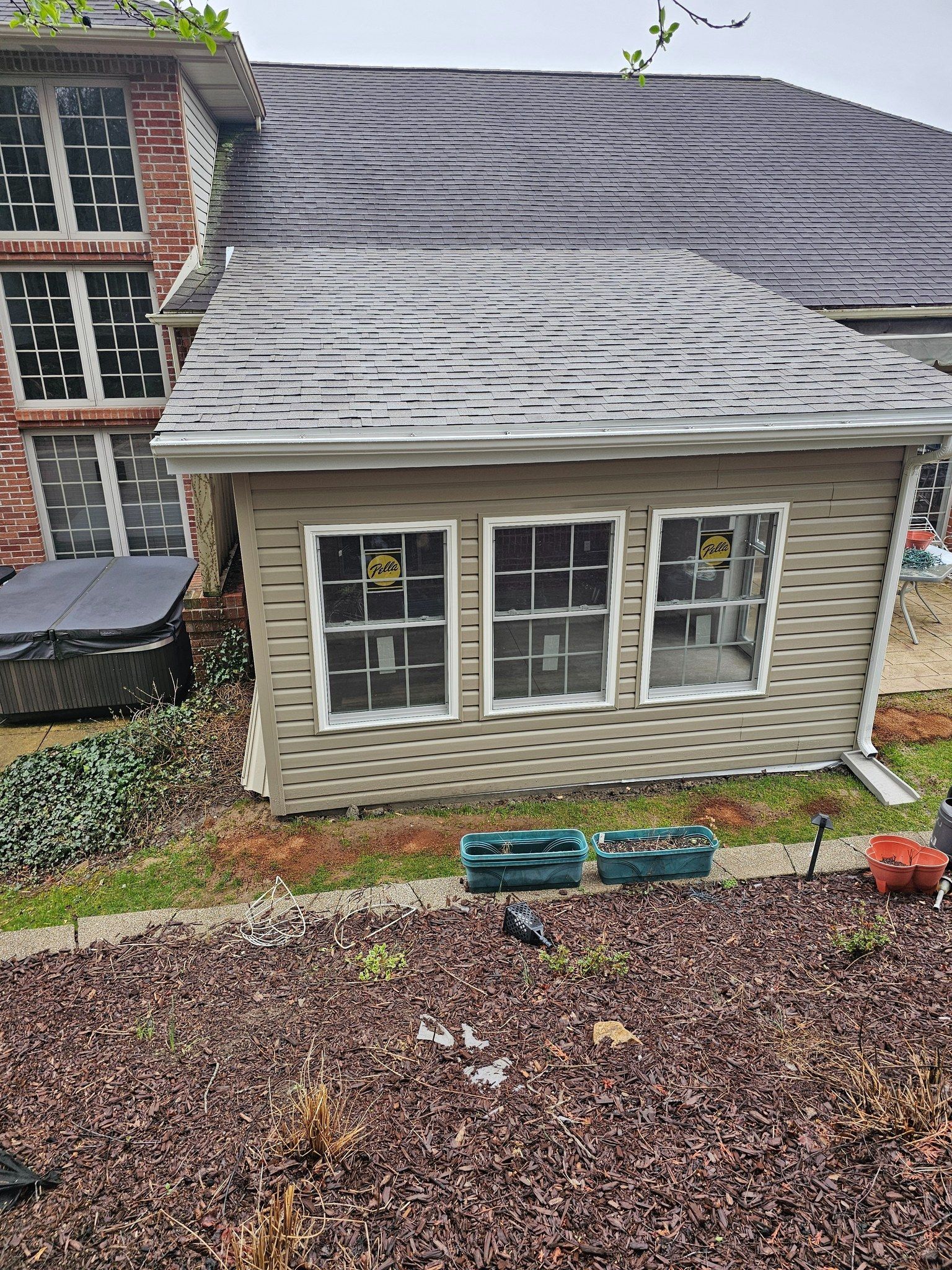 Small building with three windows, attached to a brick home with a dark shingled roof, brown mulch in foreground.