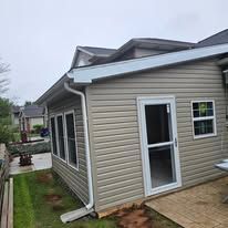 Tan vinyl-sided sunroom with white trim and a screen door, attached to a house with a green lawn.