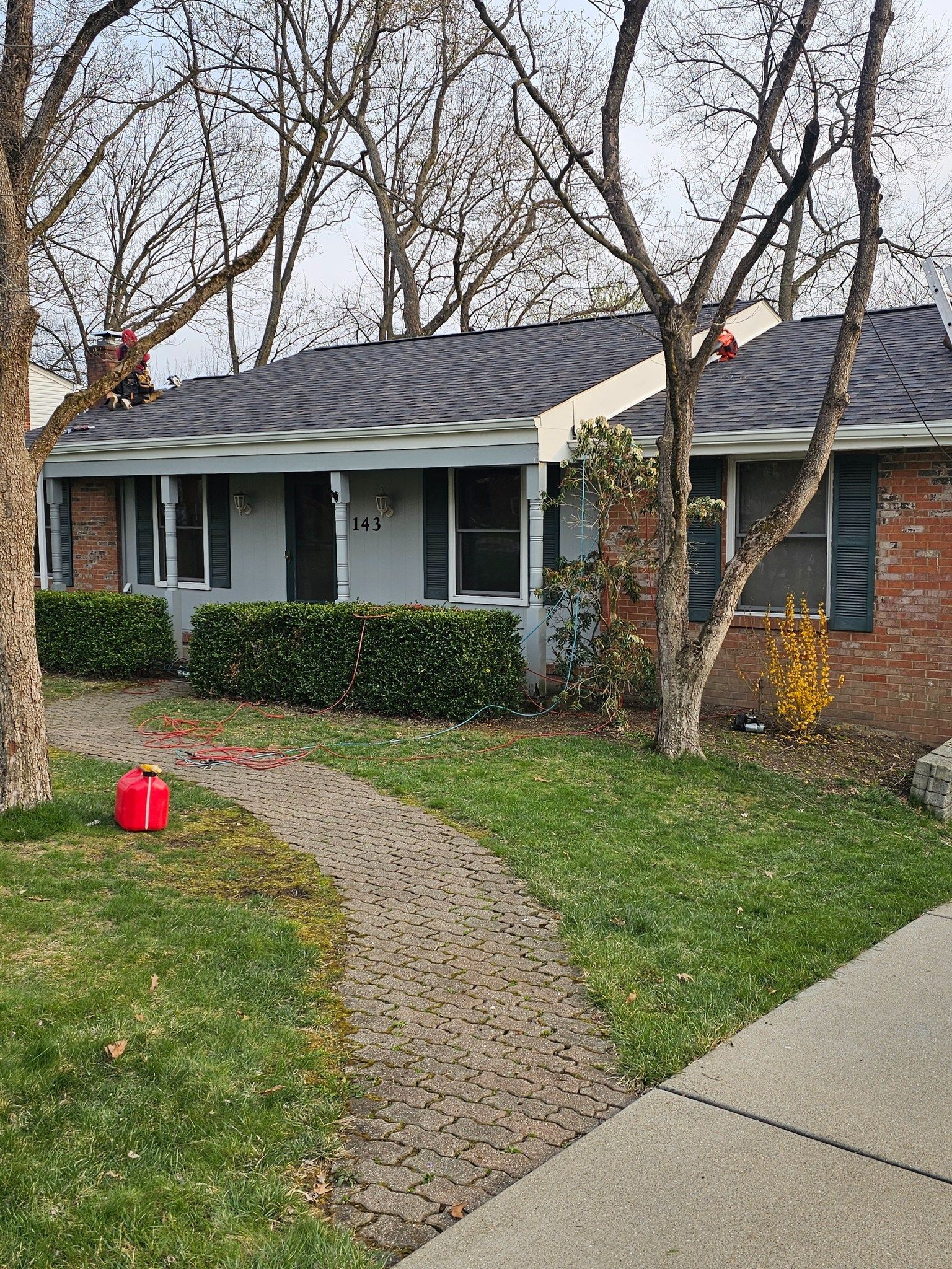 House with brick facade, hedge, pathway, trees. Green lawn, blue siding, gray roof, red can.