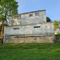 Two-story, weathered building with horizontal siding, concrete base, and several rectangular windows under a blue sky.